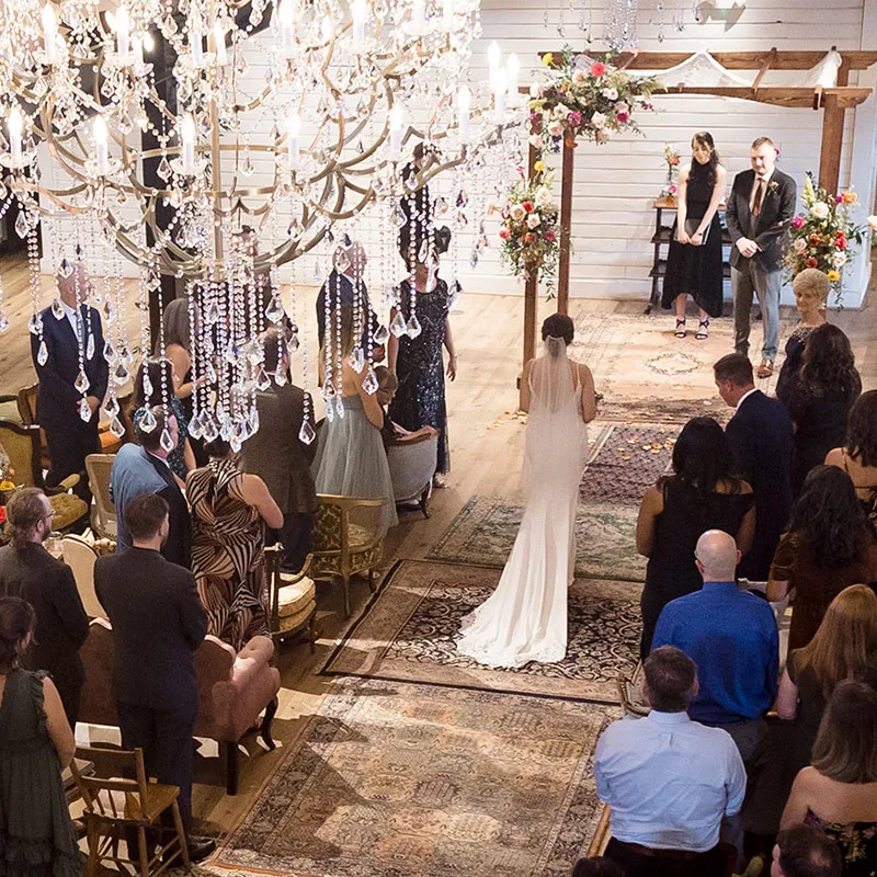 Bride walks down the carpeted aisle towards the groom who awaits with a smile amongst hanging bouquets
