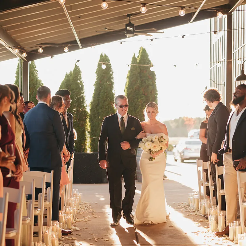 Walking down the covered outdoor aisle among white flower petals, a bride on her father's arm. Brentsville Hall, Manassas, Virginia
