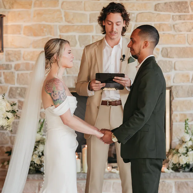 A bride smiles during the wedding ceremony next to a large stone fireplace at the covered outdoor venue. Brentsville Hall, Manassas, Virginia.
