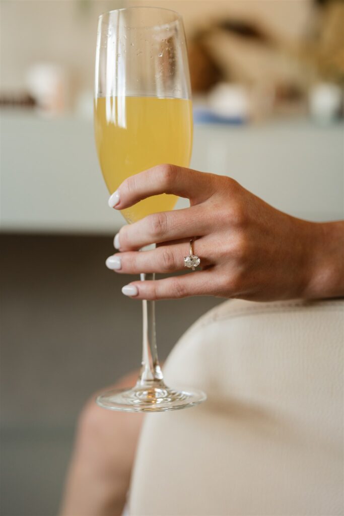 A bride prepares for her wedding with a mimosa and hand, her engagement ring on display